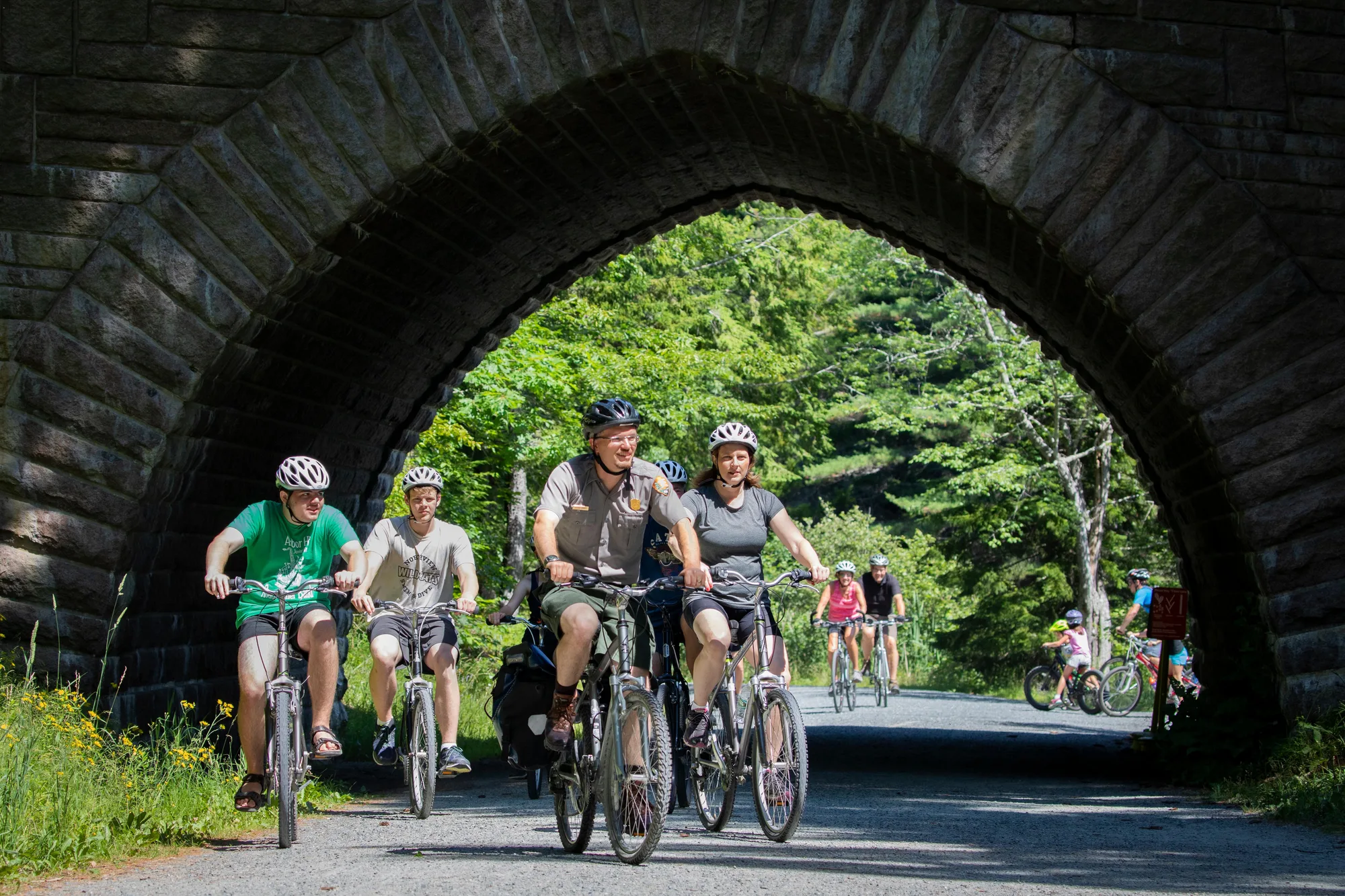A family rides bicycles under a stone carriage-road arch in Acadia National Park